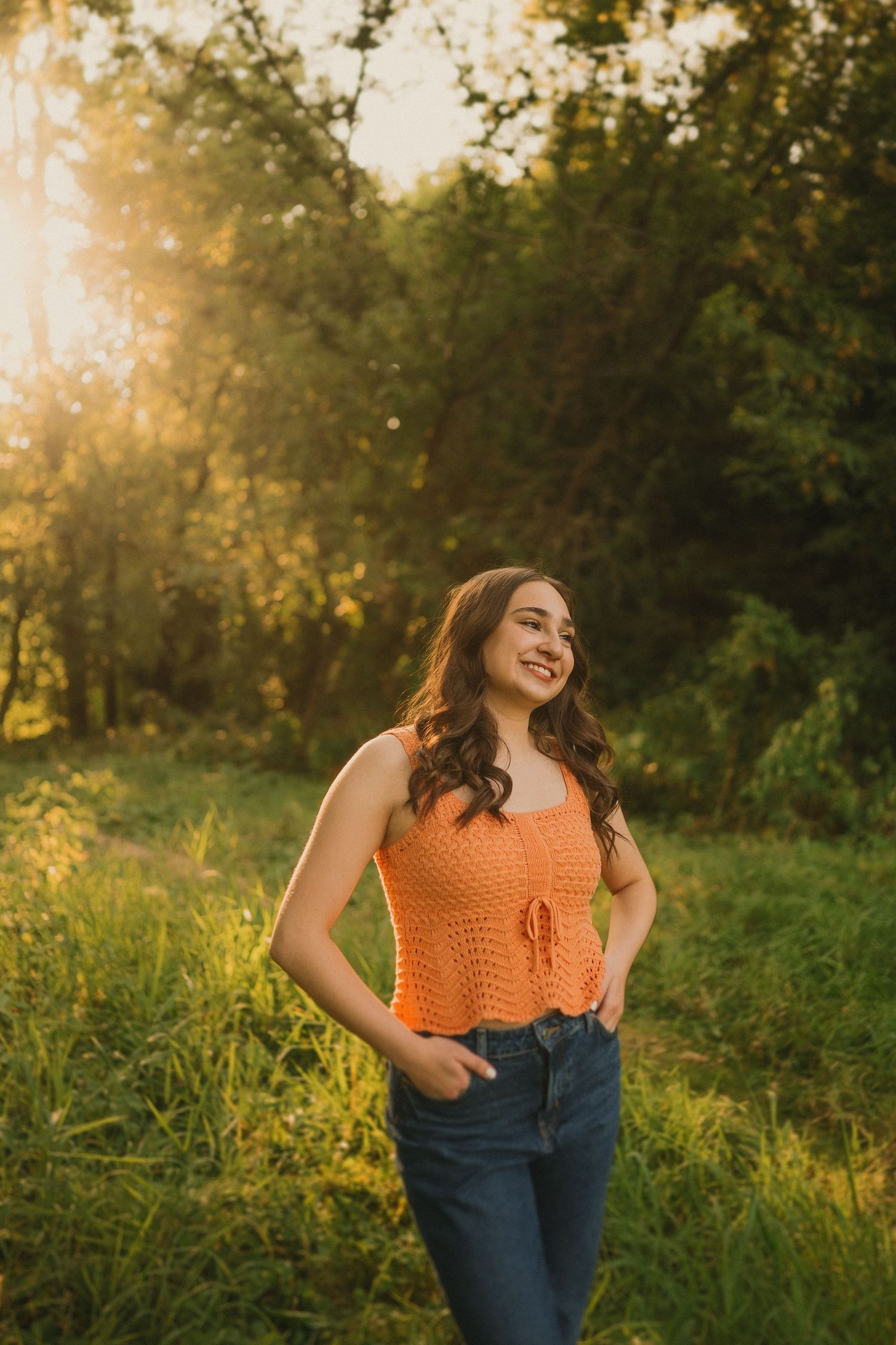 A summer portrait in an orange crochet top and jeans standing in a sun-drenched field at sunset.