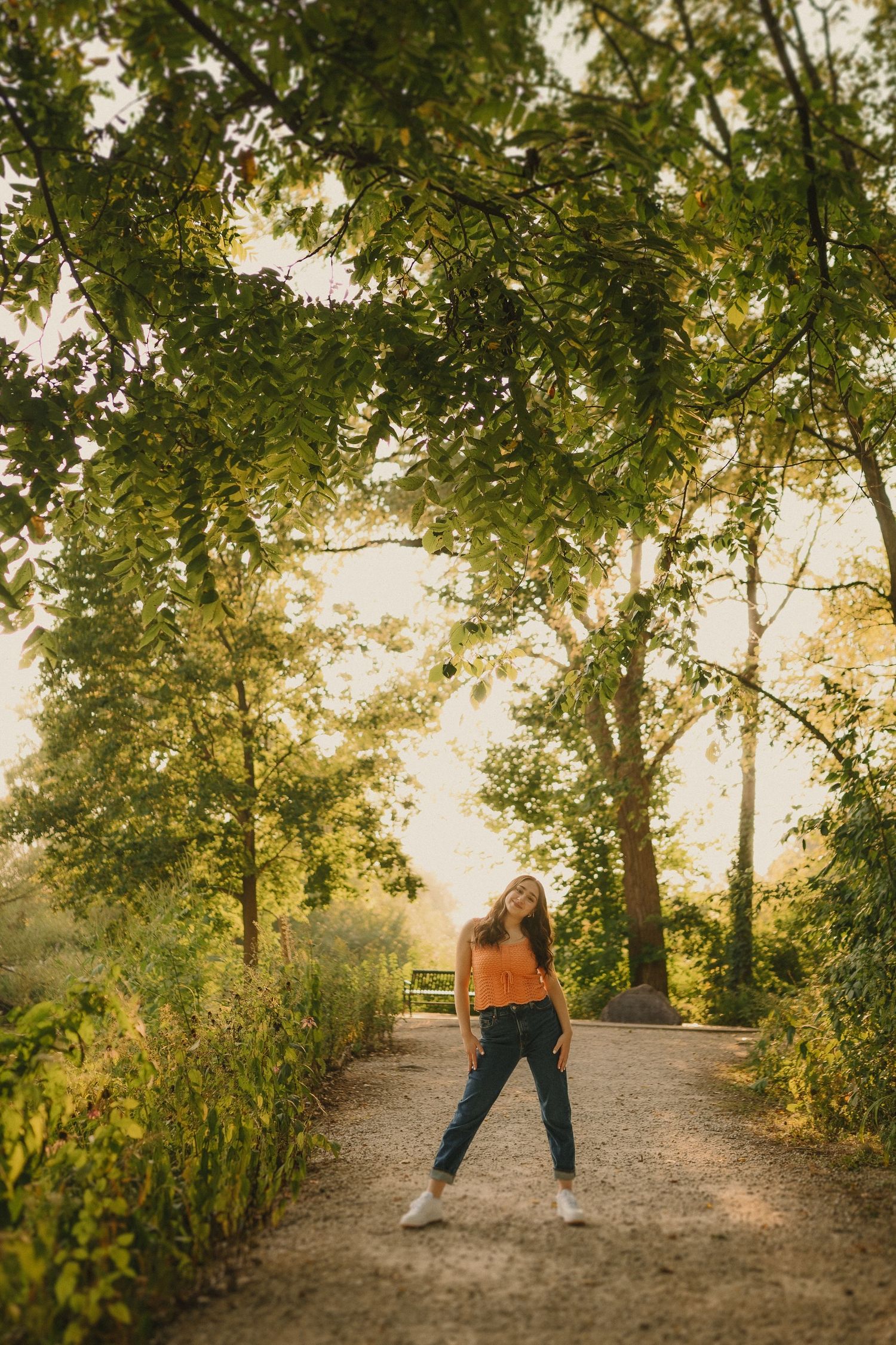 A sunlit dirt path through green trees with a figure standing in casual autumn attire creating a warm, scenic atmosphere.