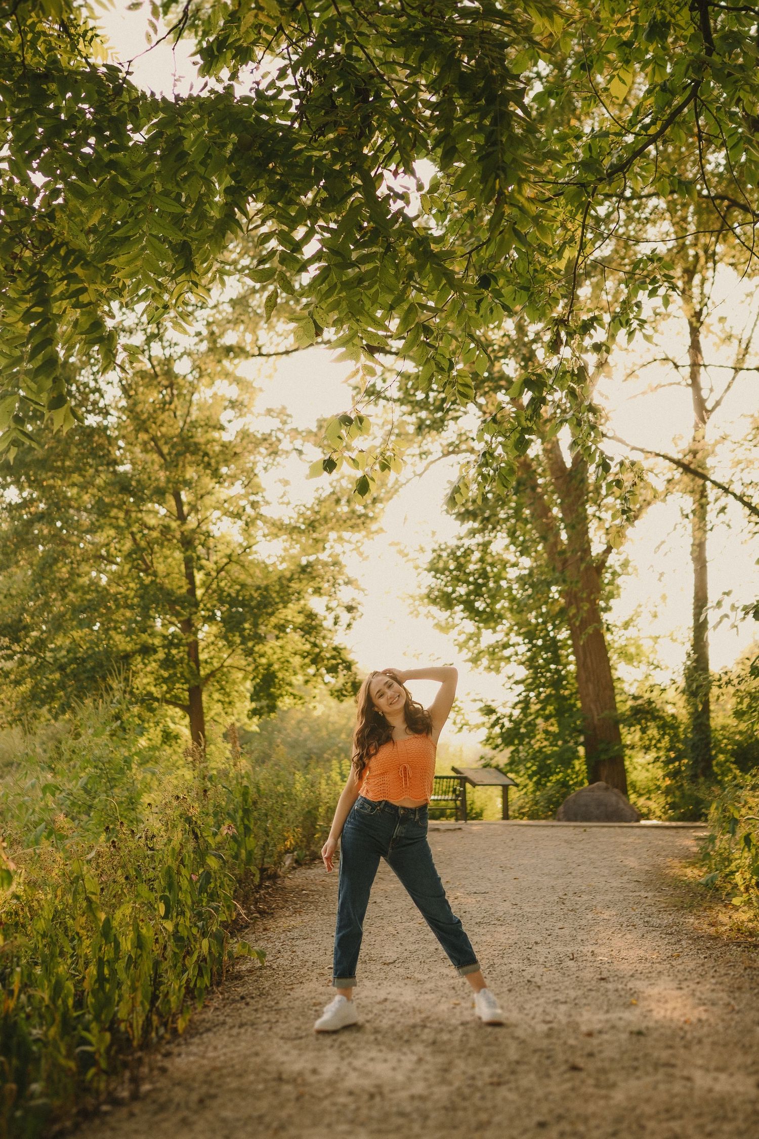 Person in an orange top and jeans strikes a playful pose on a tree-lined path during golden hour.