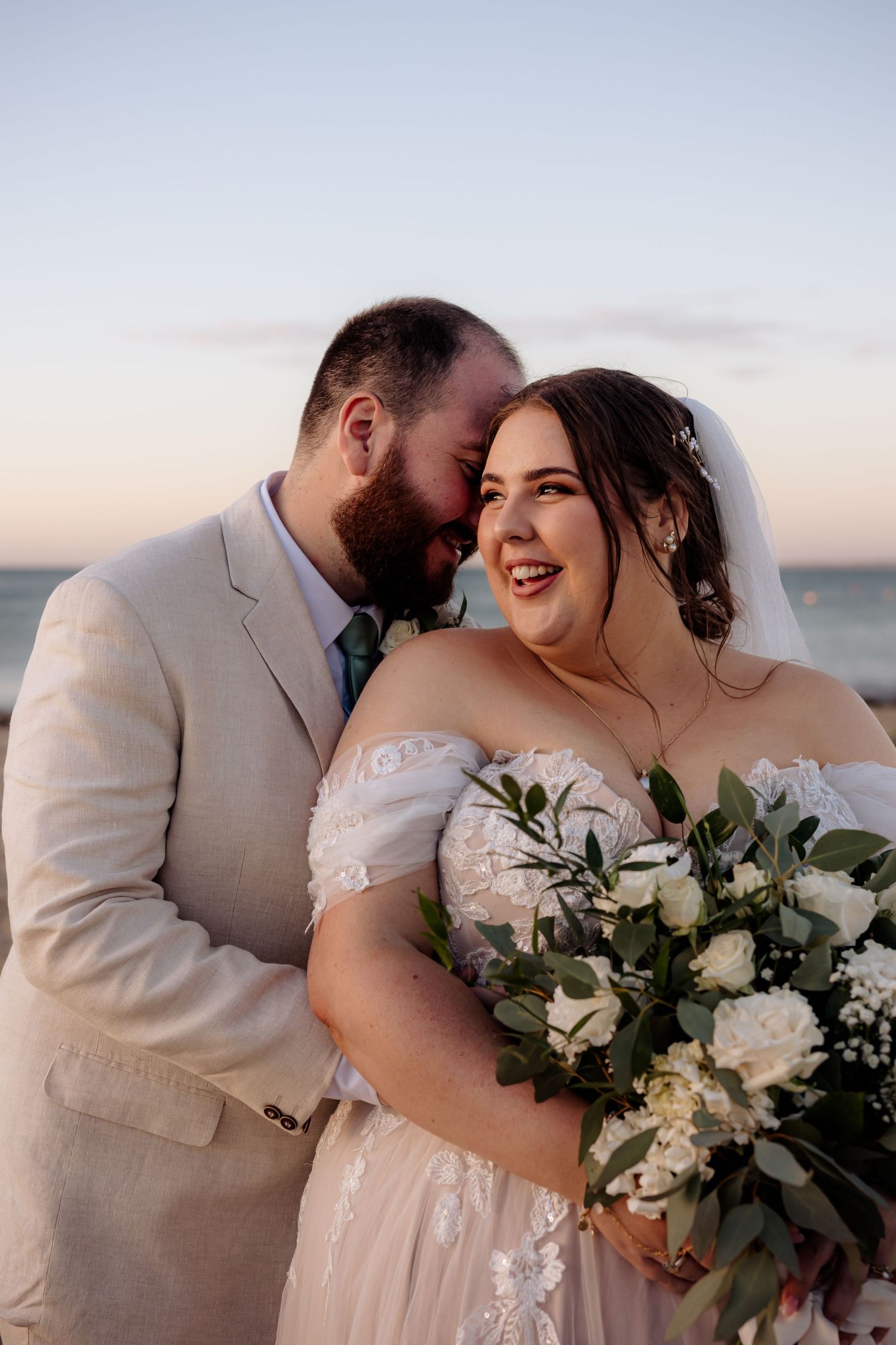 A romantic beach wedding couple shares a tender moment at sunset with a beautiful white and green floral bouquet.