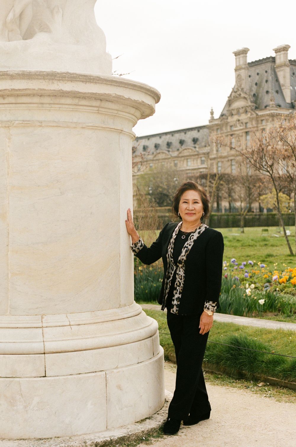 A person in a black outfit poses beside a white marble column in the Tuileries Garden with the Louvre Museum visible in background.