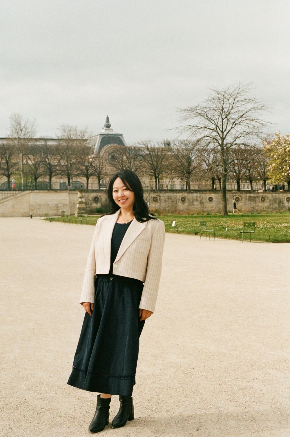 A person in a cream blazer and black dress poses at a garden path with historic architecture in the background.