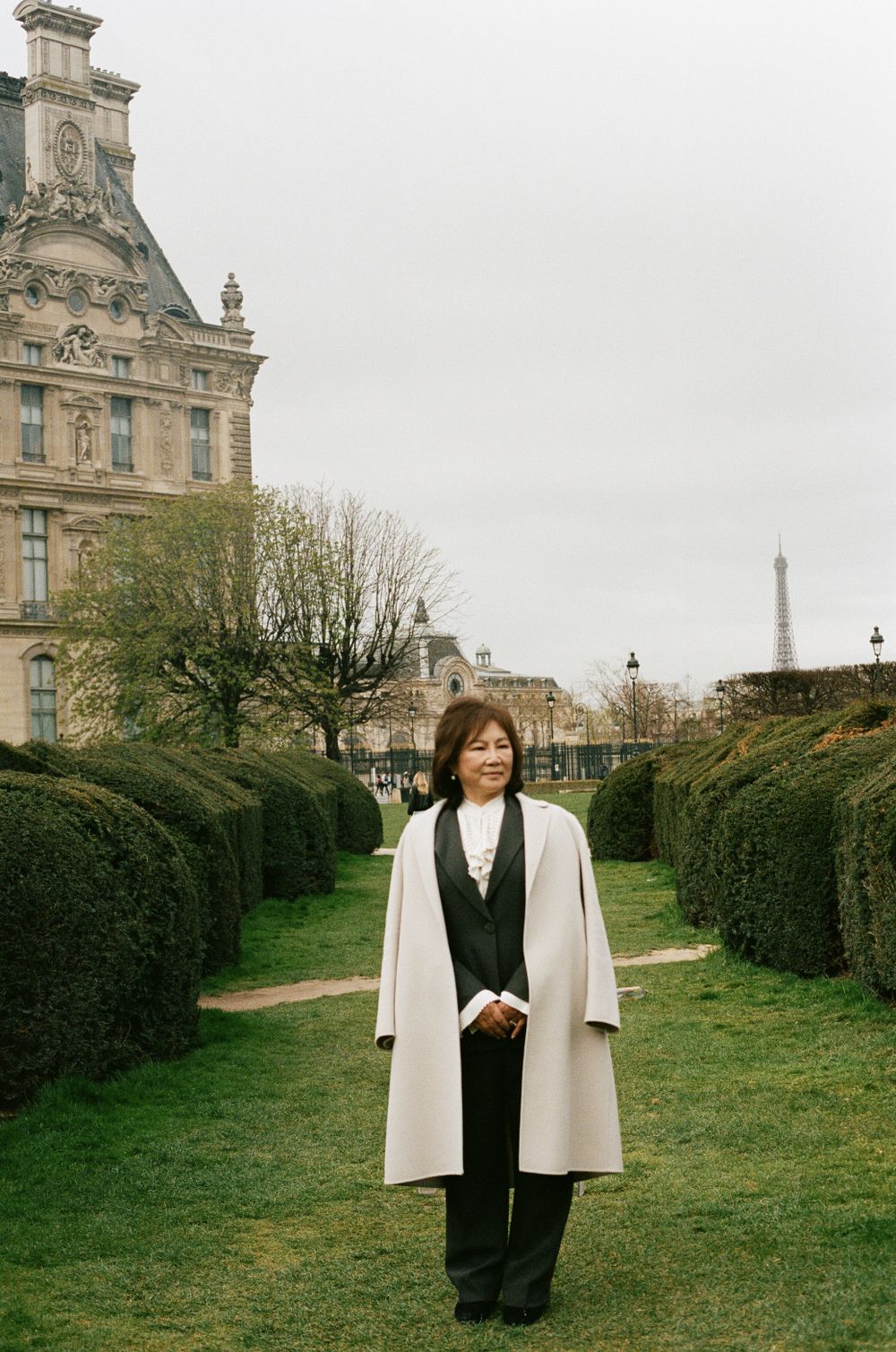 A person in a white coat and black outfit stands in the Tuileries Garden in Paris with the Eiffel Tower visible in the distance.