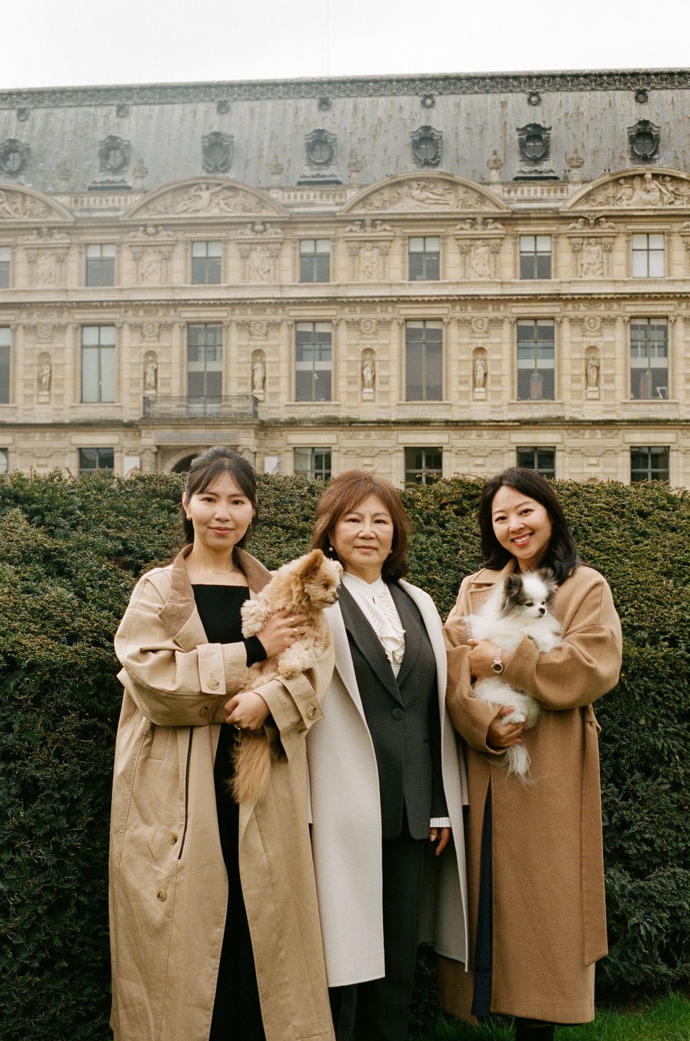 Three people in winter coats posing together in front of an ornate European palace building with manicured hedges.