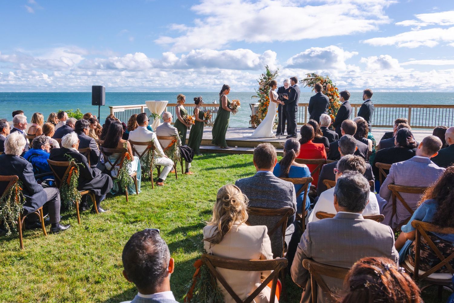 A beautiful outdoor wedding ceremony takes place on a grassy cliff overlooking the bright blue ocean on a sunny day.