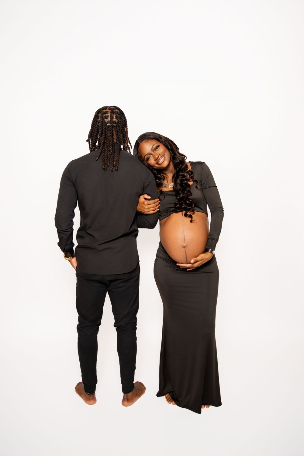 Couple in black outfits poses for casual maternity photo against white background.