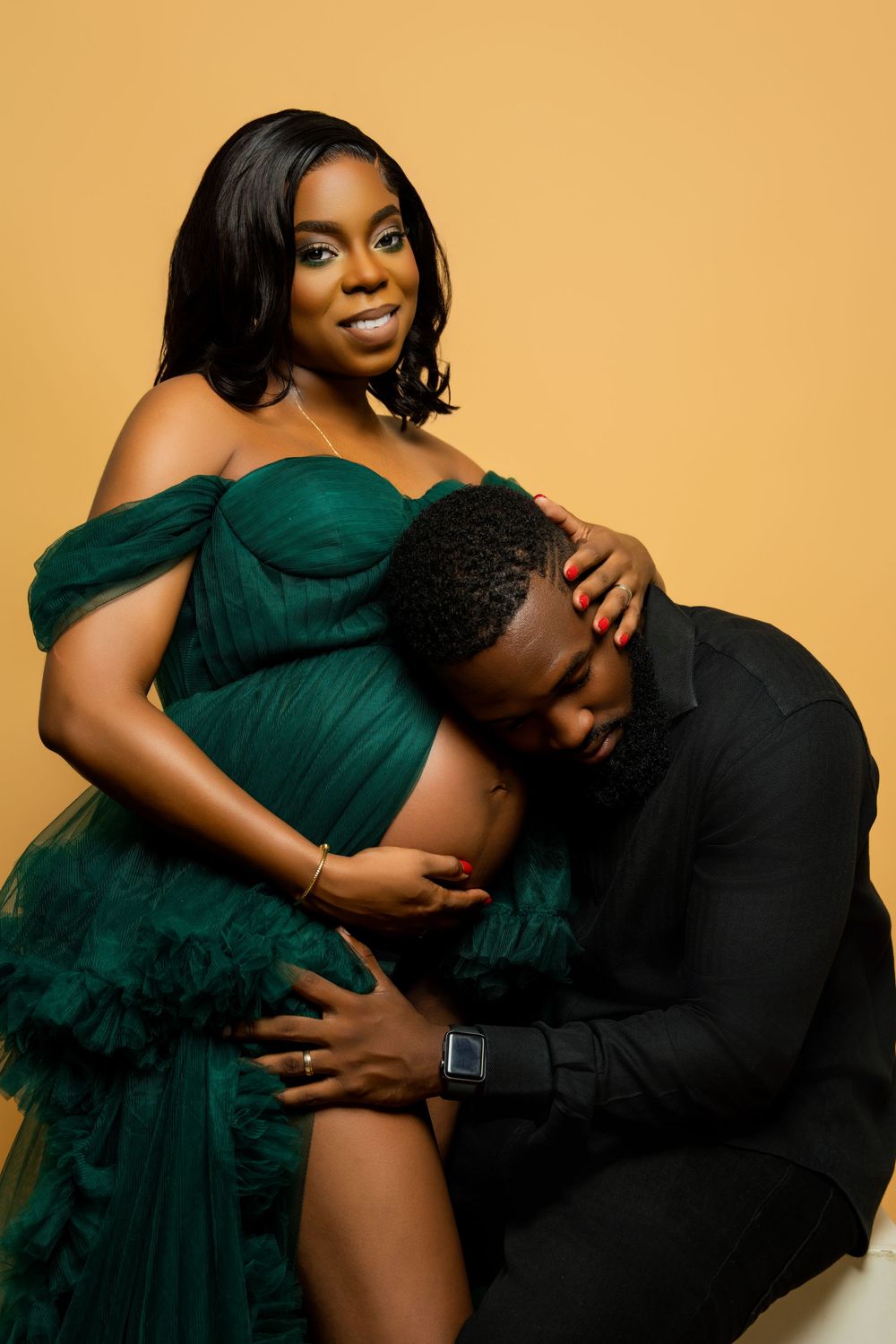 Couple in formal wear share intimate maternity moment against golden background.