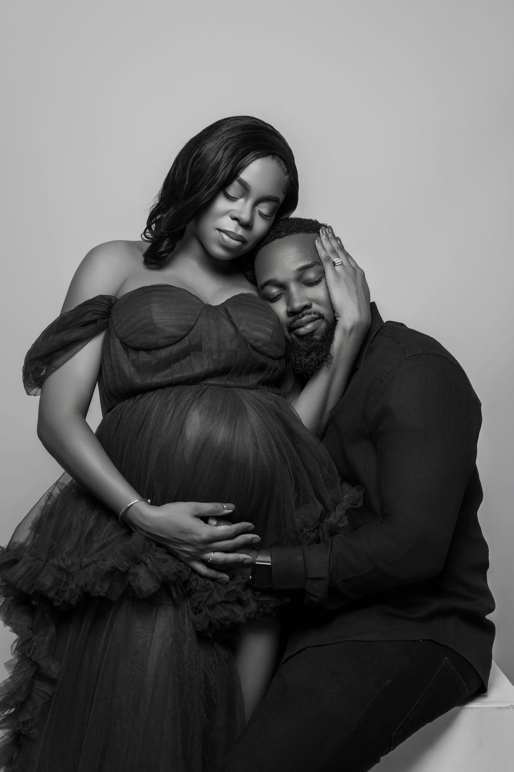 Intimate black and white portrait of an expectant couple embracing tenderly against a neutral background.