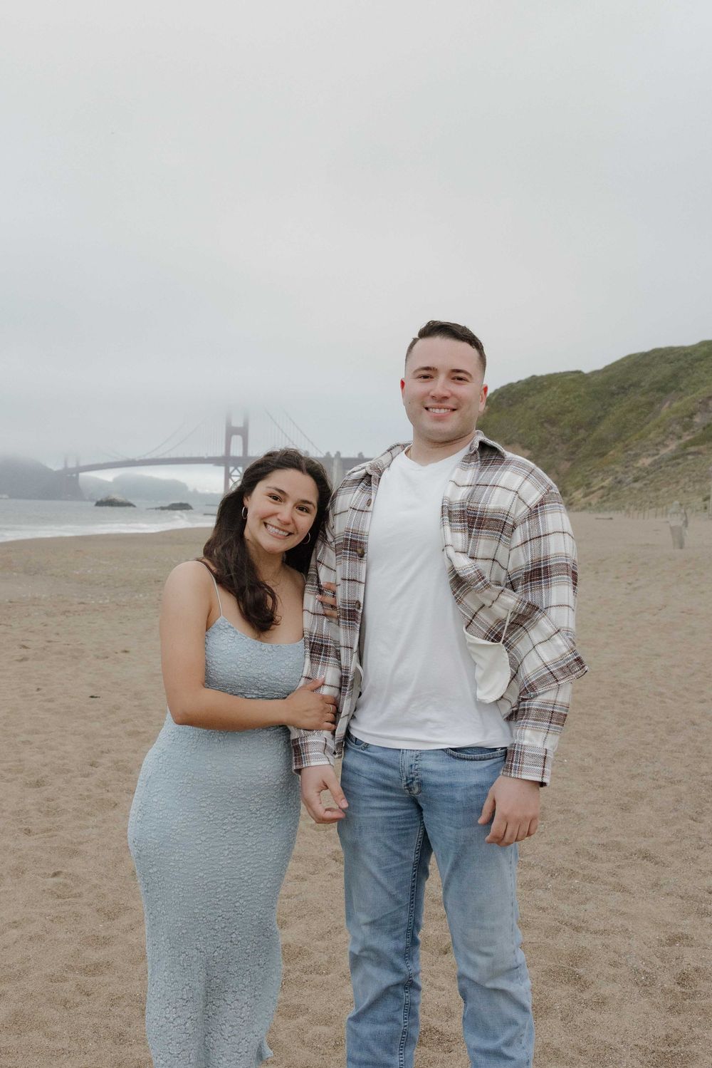 A couple stands together on a beach with the Golden Gate Bridge visible in the foggy background.