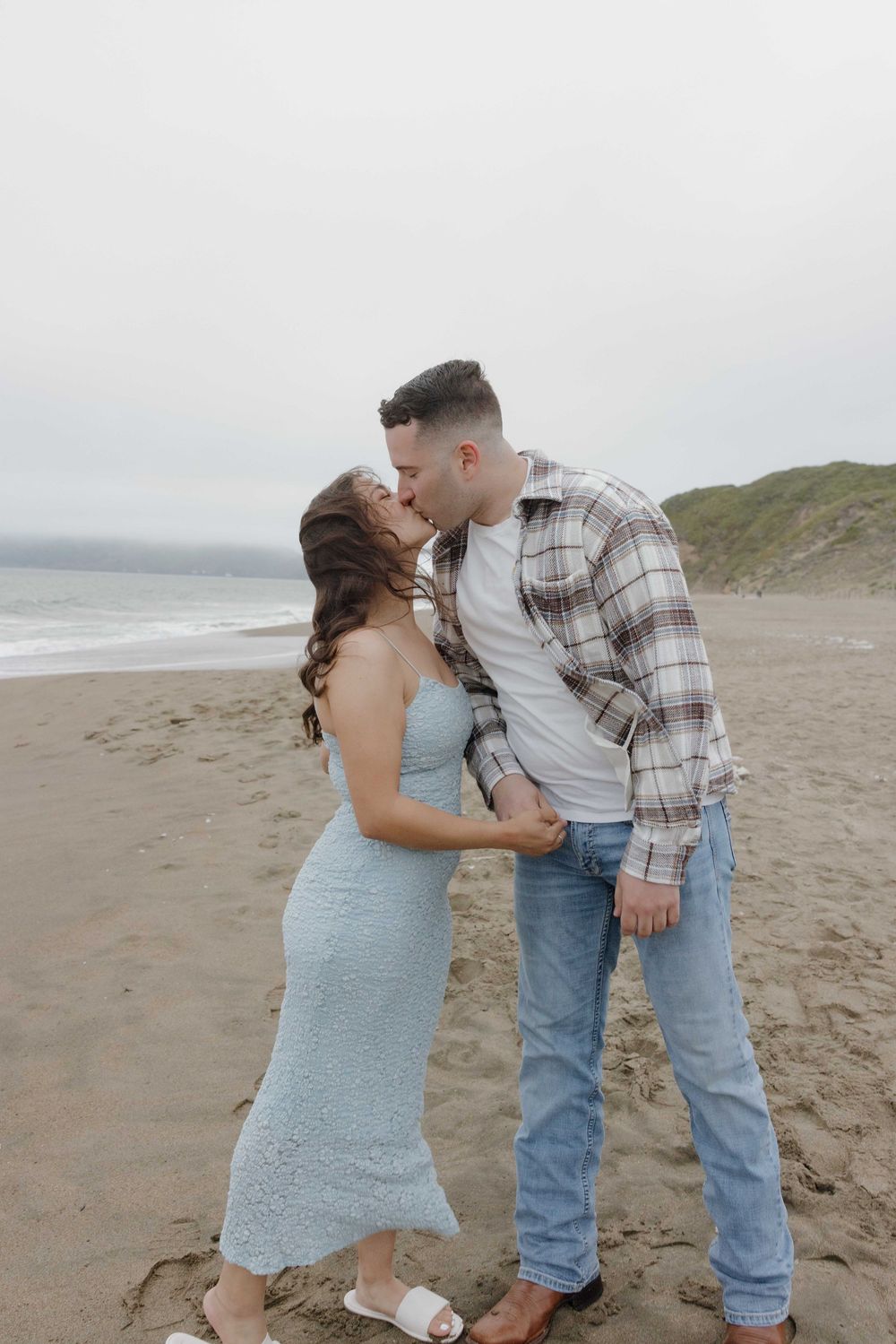 A romantic beach scene with a couple sharing a tender kiss on a foggy coastline near the ocean.
