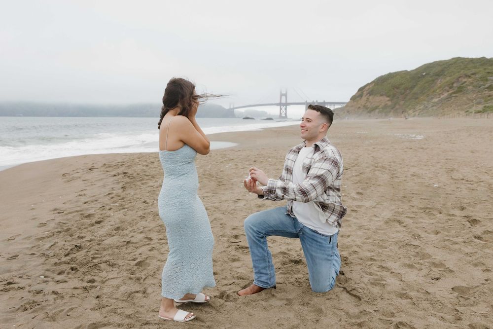 A romantic proposal scene unfolds on a misty beach with coastal cliffs and bridge in the background.
