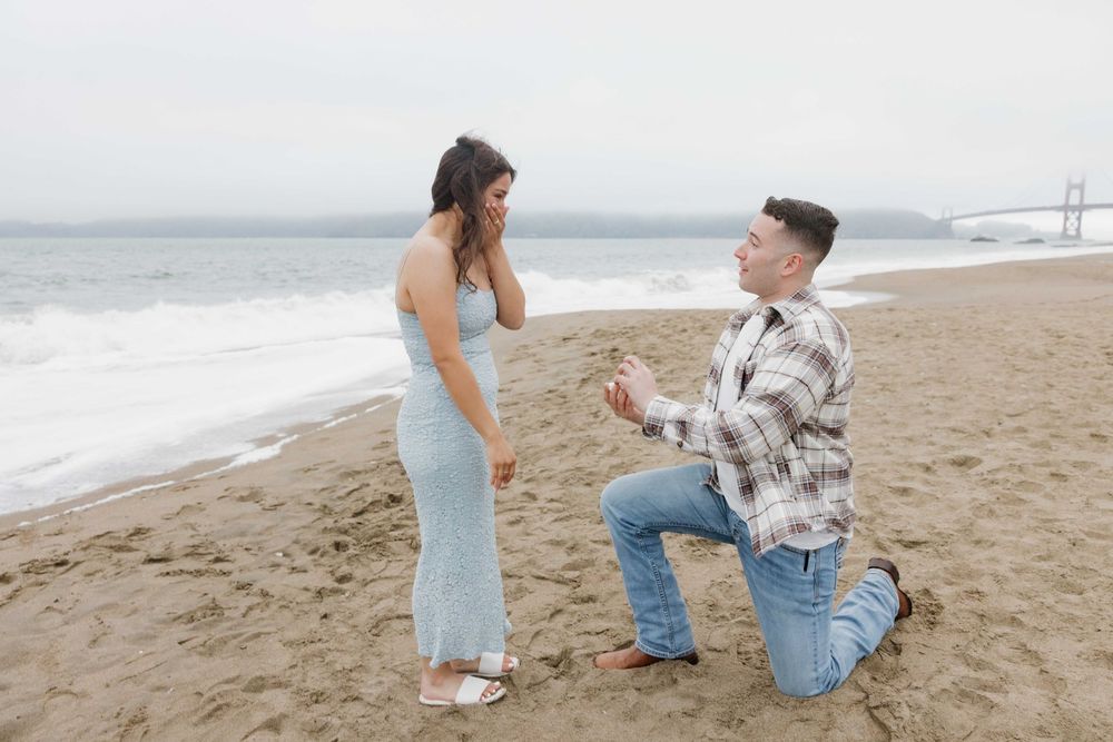 A person proposes on one knee to their partner on a cloudy beach near the ocean.