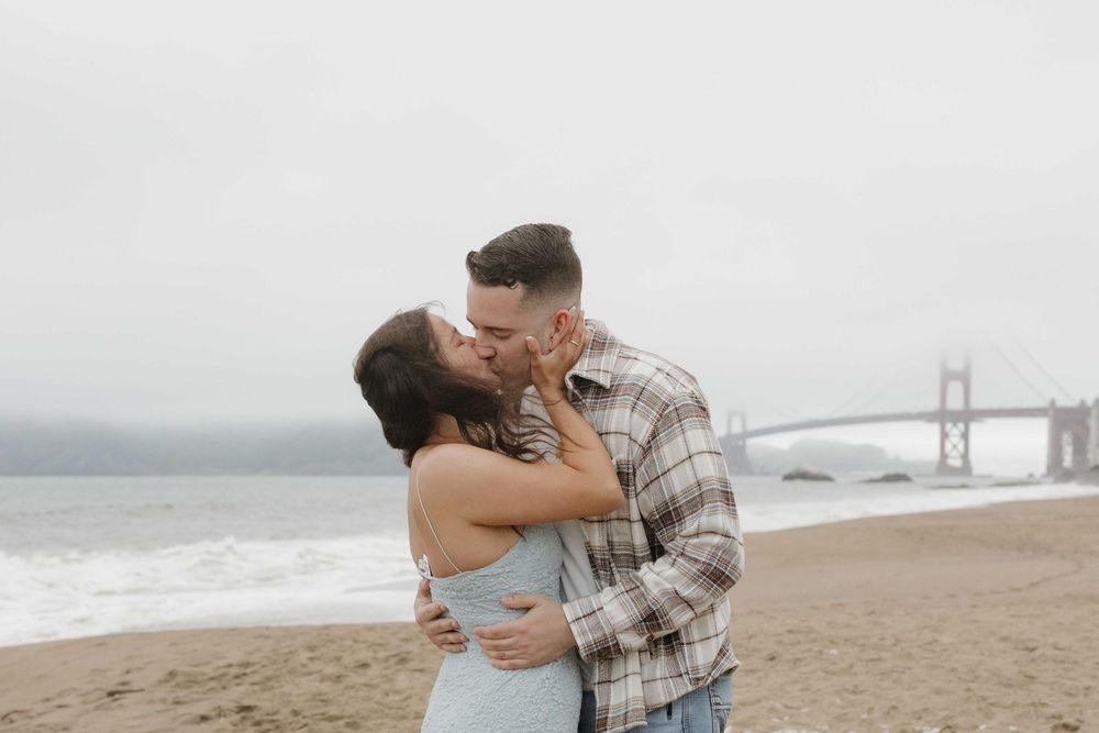 A romantic moment shared between a couple embracing on Baker Beach with the Golden Gate Bridge in the background.