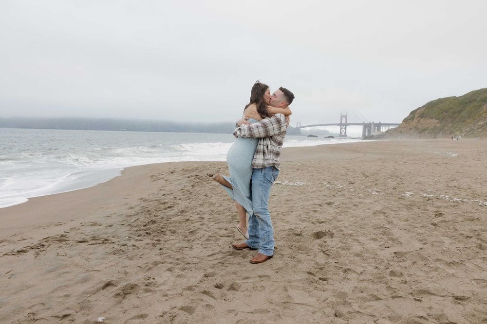 A couple sharing an intimate moment on a foggy beach with the Golden Gate Bridge visible in the distance.
