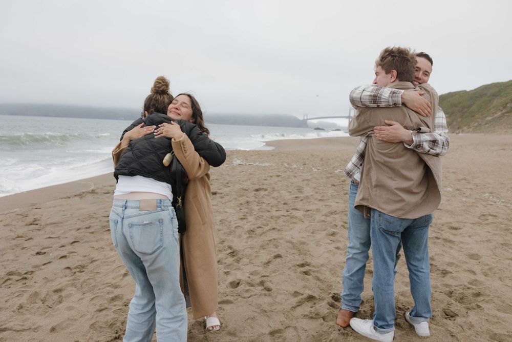 Two couples embrace each other on a foggy beach with rolling waves and hills in the background.
