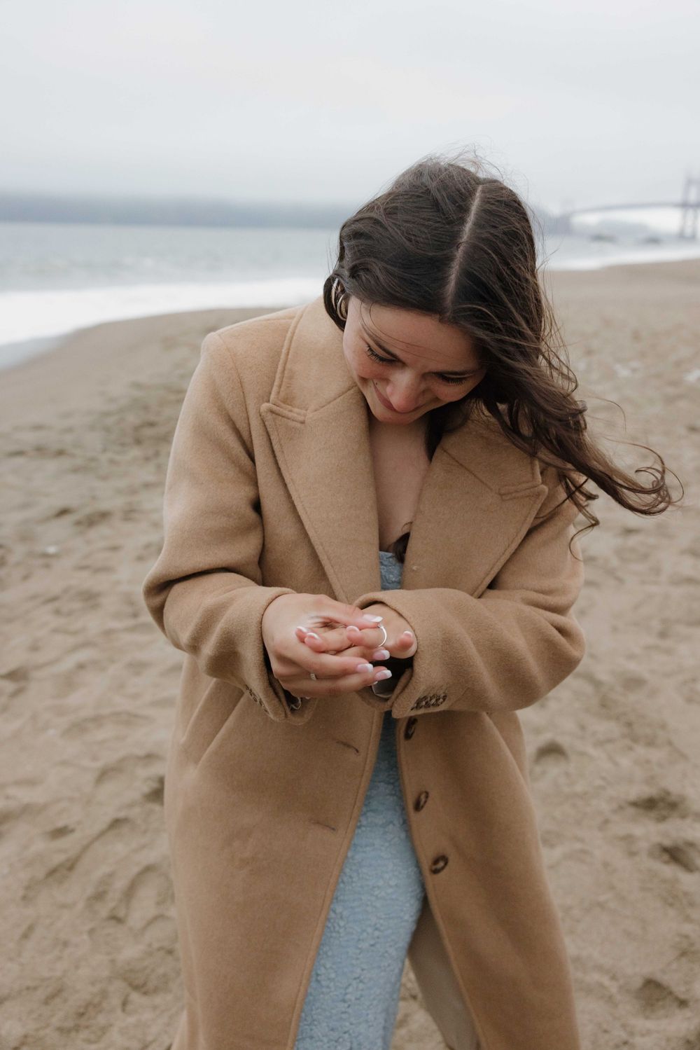 Person wearing a camel coat smiles warmly on a foggy beach while looking down at hands.