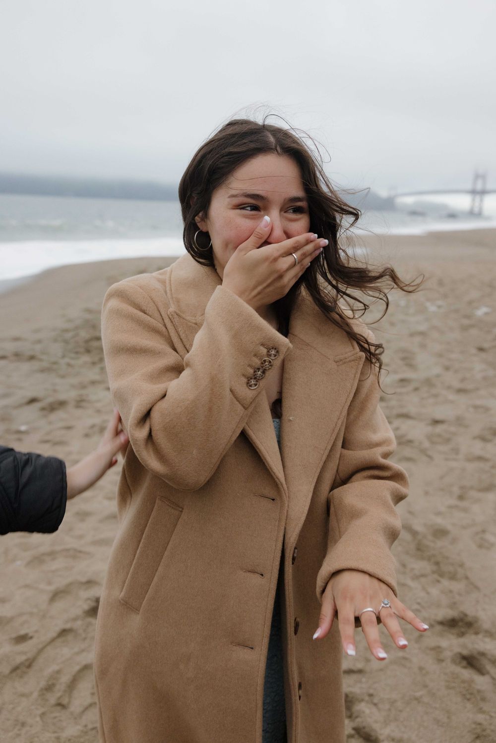 Someone wearing a camel coat laughs with hand covering mouth while standing on a foggy beach.