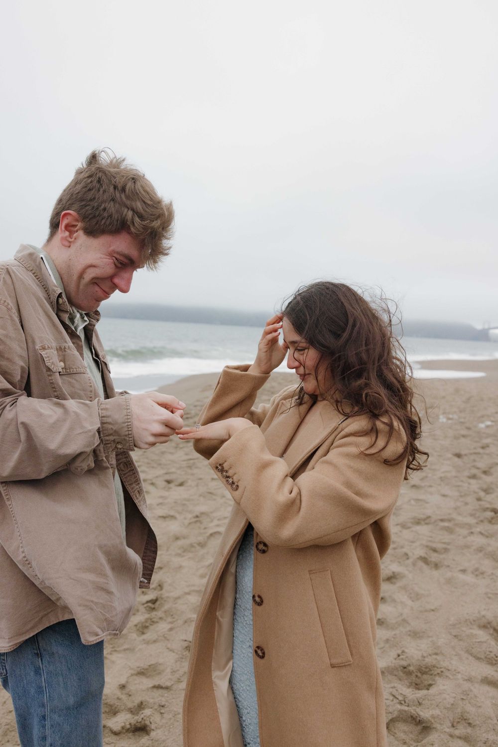 Two people sharing a joyful moment while standing on a cloudy beach with one wearing a tan coat and one in brown jacket.