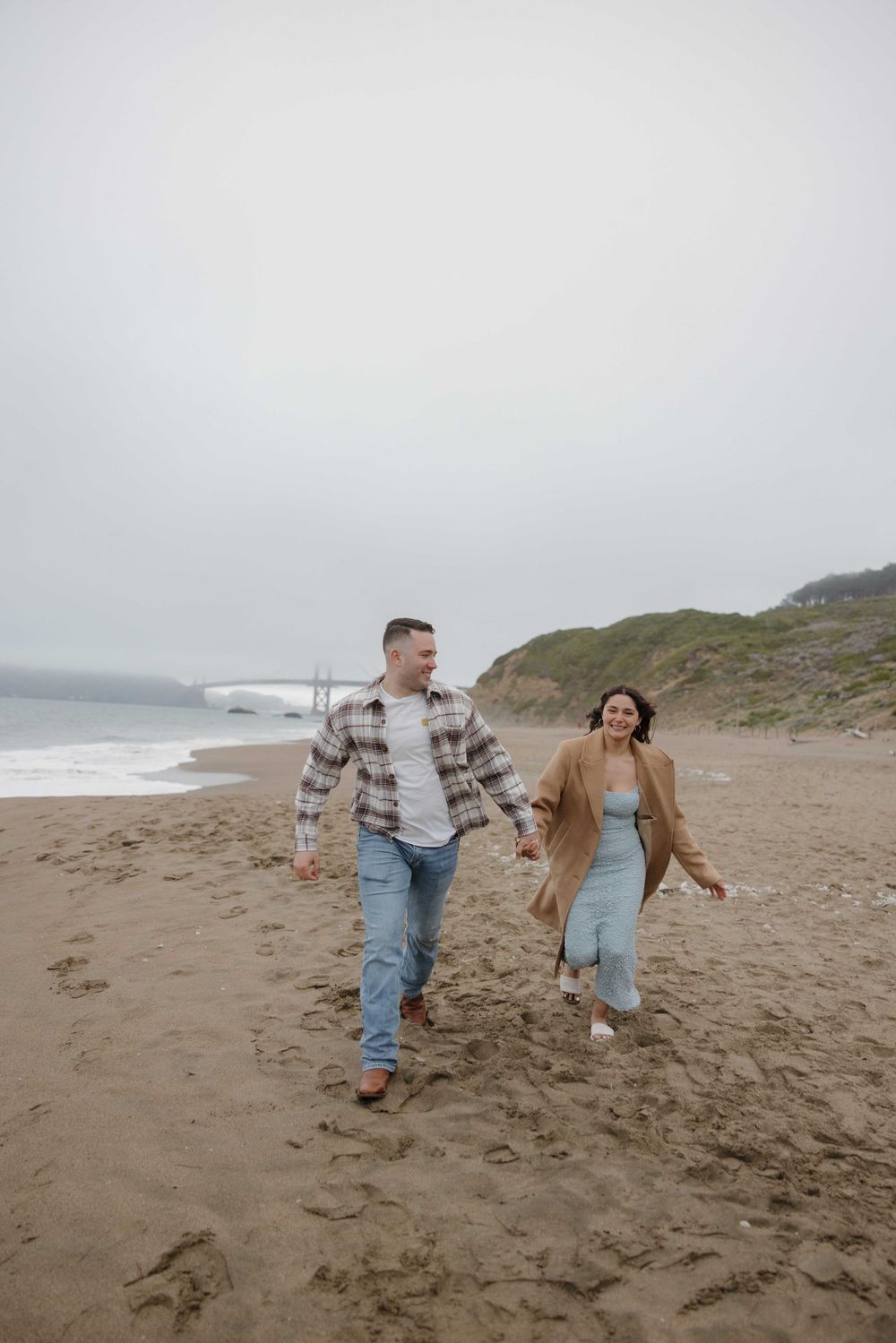 A couple walks and smiles together on a misty beach with person in plaid shirt and person in blue dress.