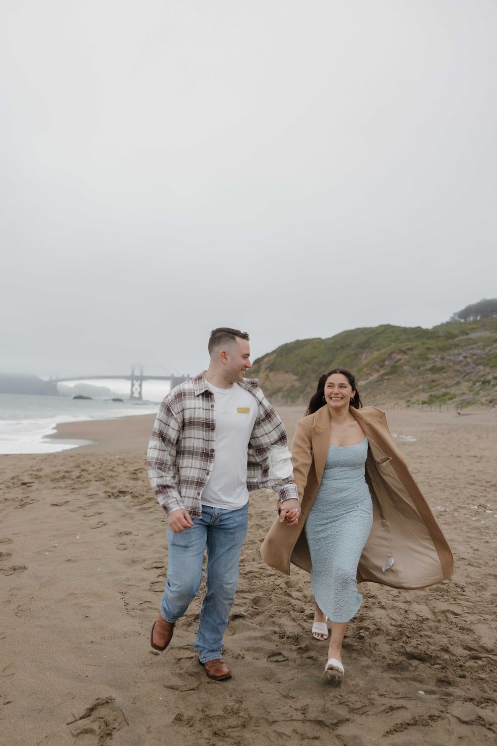 A couple walking joyfully along a foggy beach with cliffs and bridge in the distance.