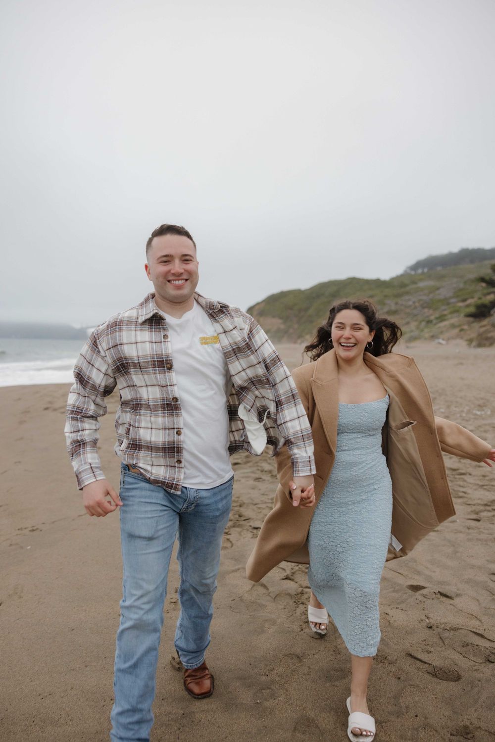 A couple walks joyfully along a misty beach coastline with hills in the background.
