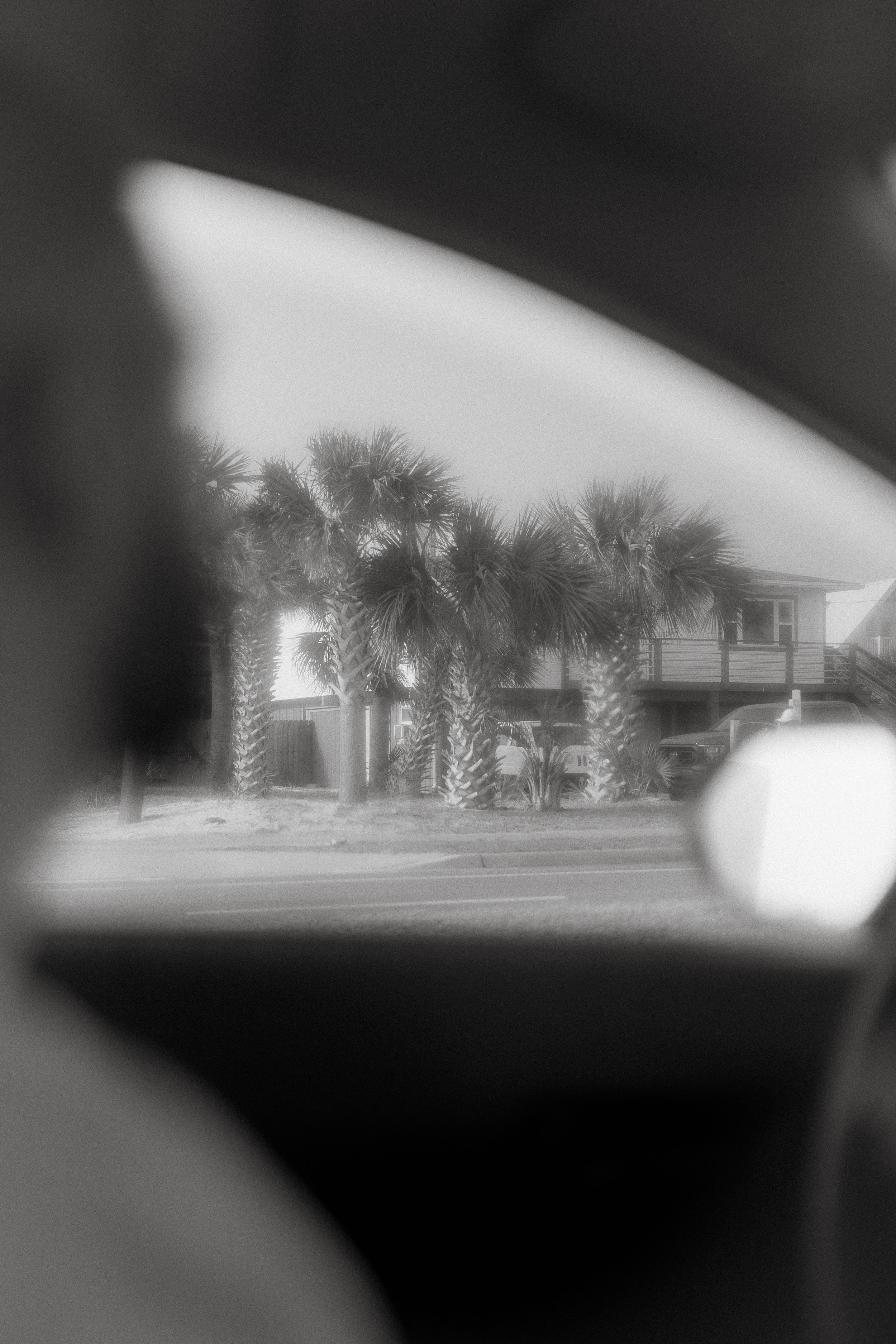 Black and white view through a car window of a beachfront house with palm trees and a porch, giving a dreamy vignette effect.
