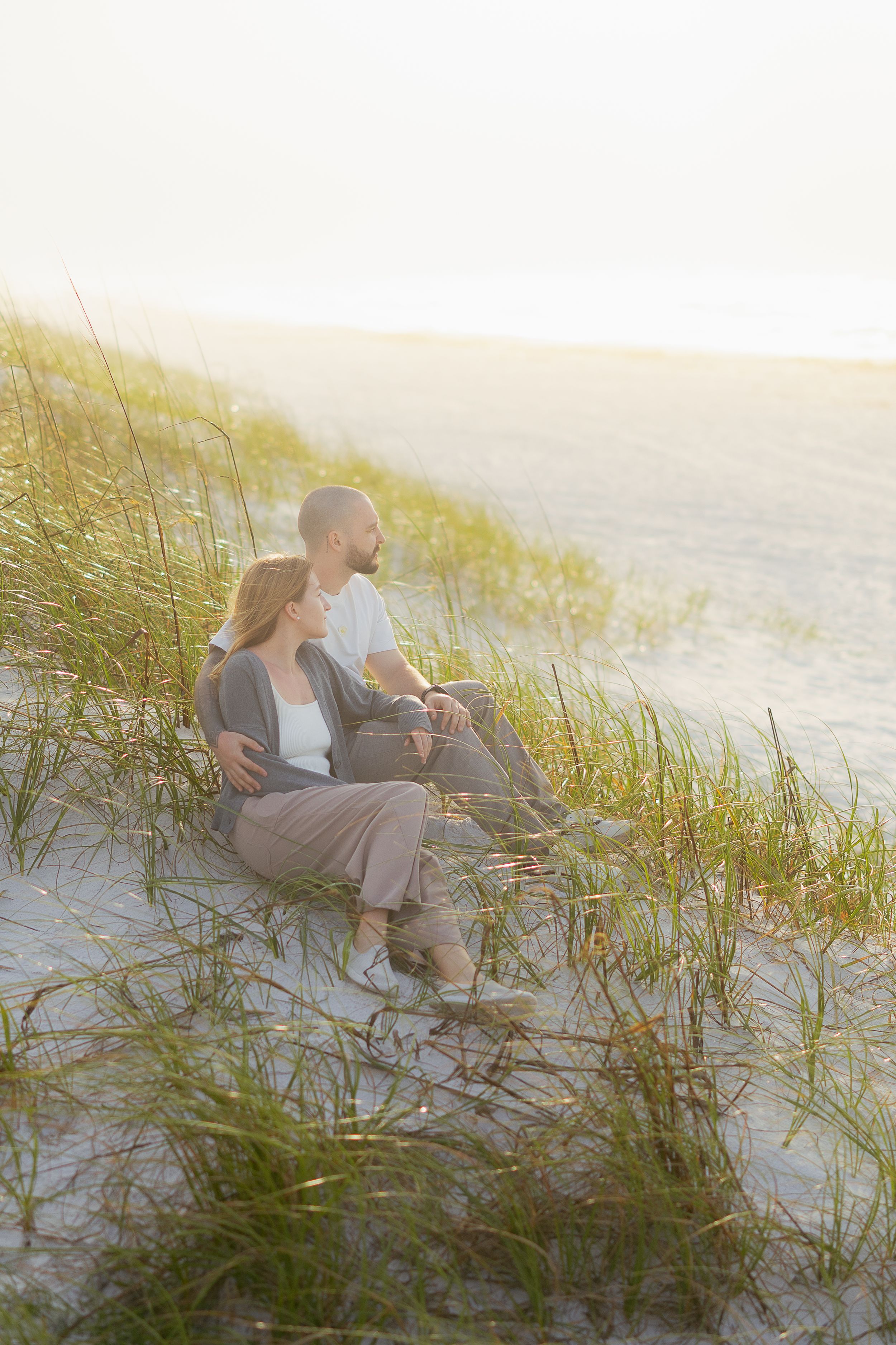 A couple sits together on sandy dunes at sunset, surrounded by beach grass and overlooking the ocean.