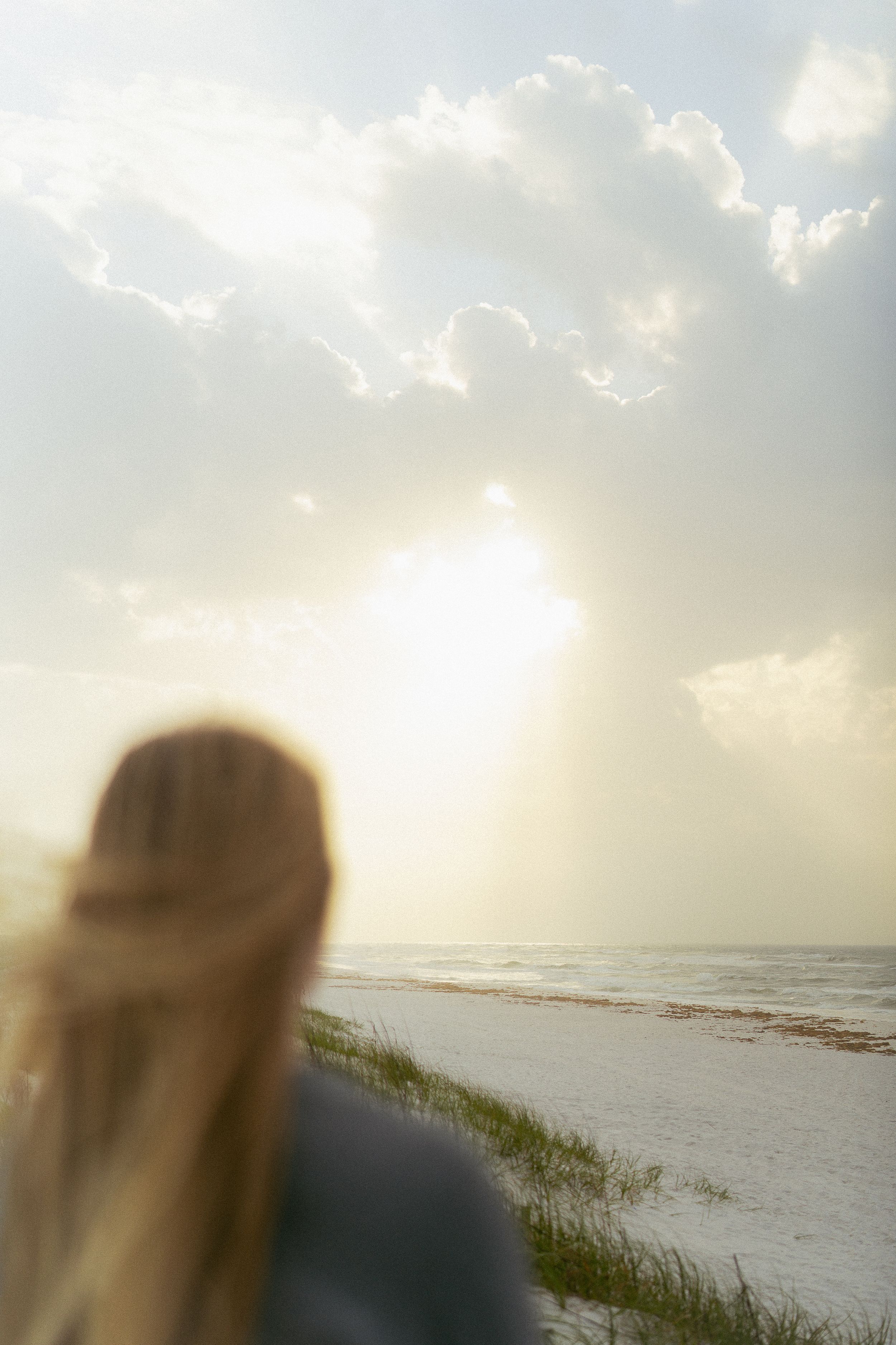Someone gazes at a peaceful beach sunset through wispy clouds as sun rays illuminate the shoreline and sea grass.