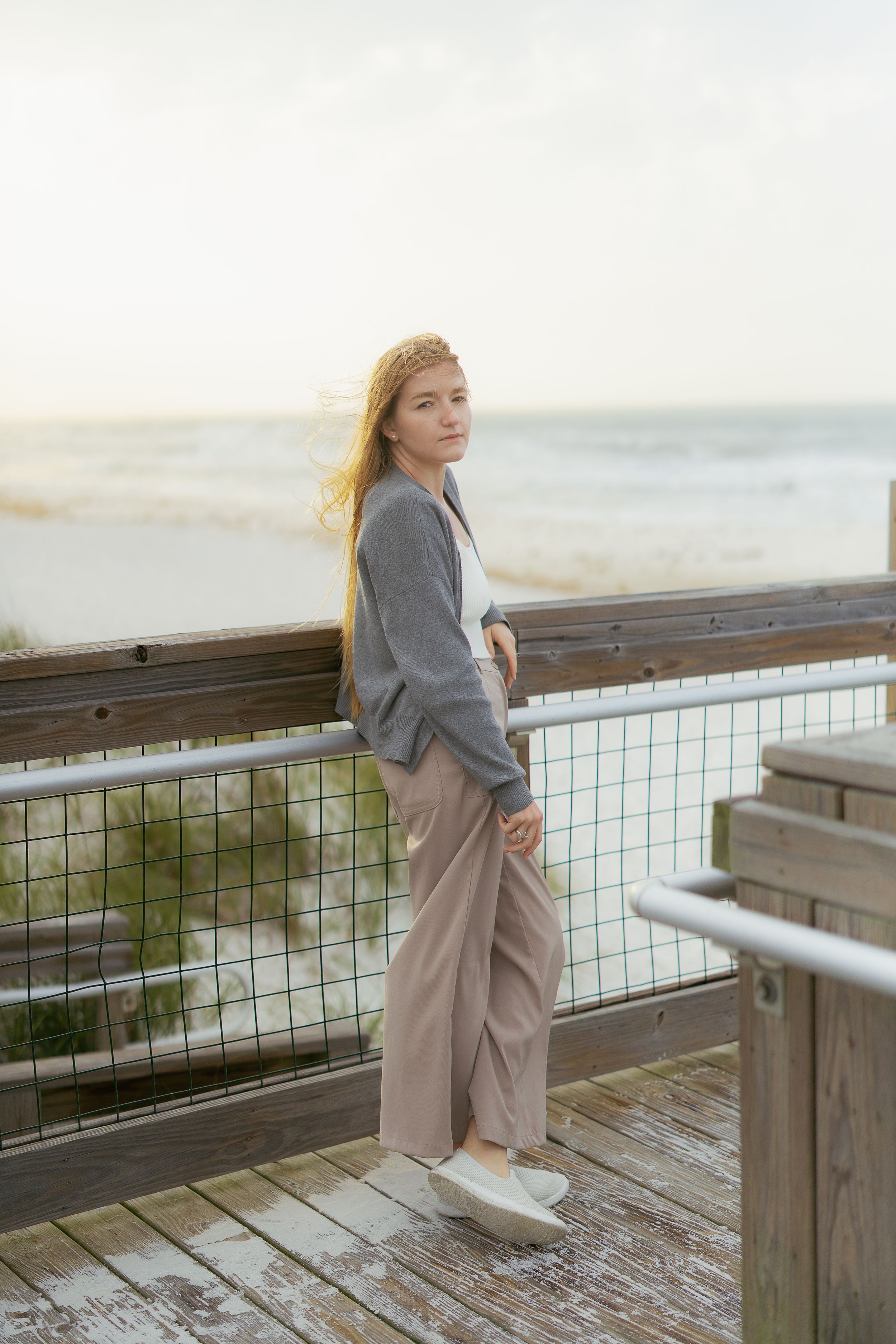Person in gray sweater and beige pants standing on wooden boardwalk overlooking coastal scenery at sunset.