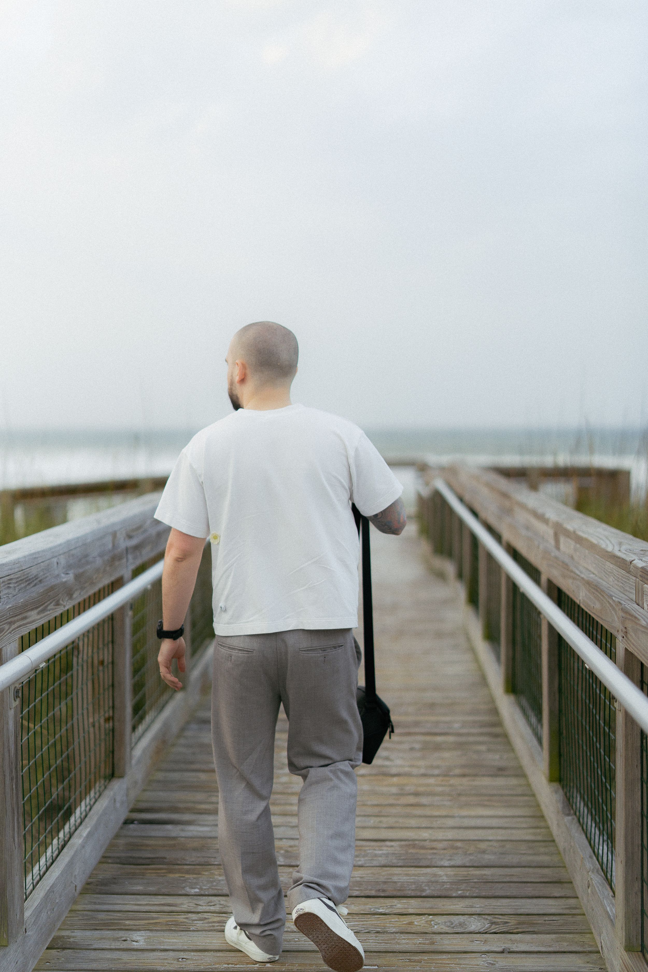 Person in white t-shirt and gray pants walking alone on a wooden boardwalk on an overcast day.