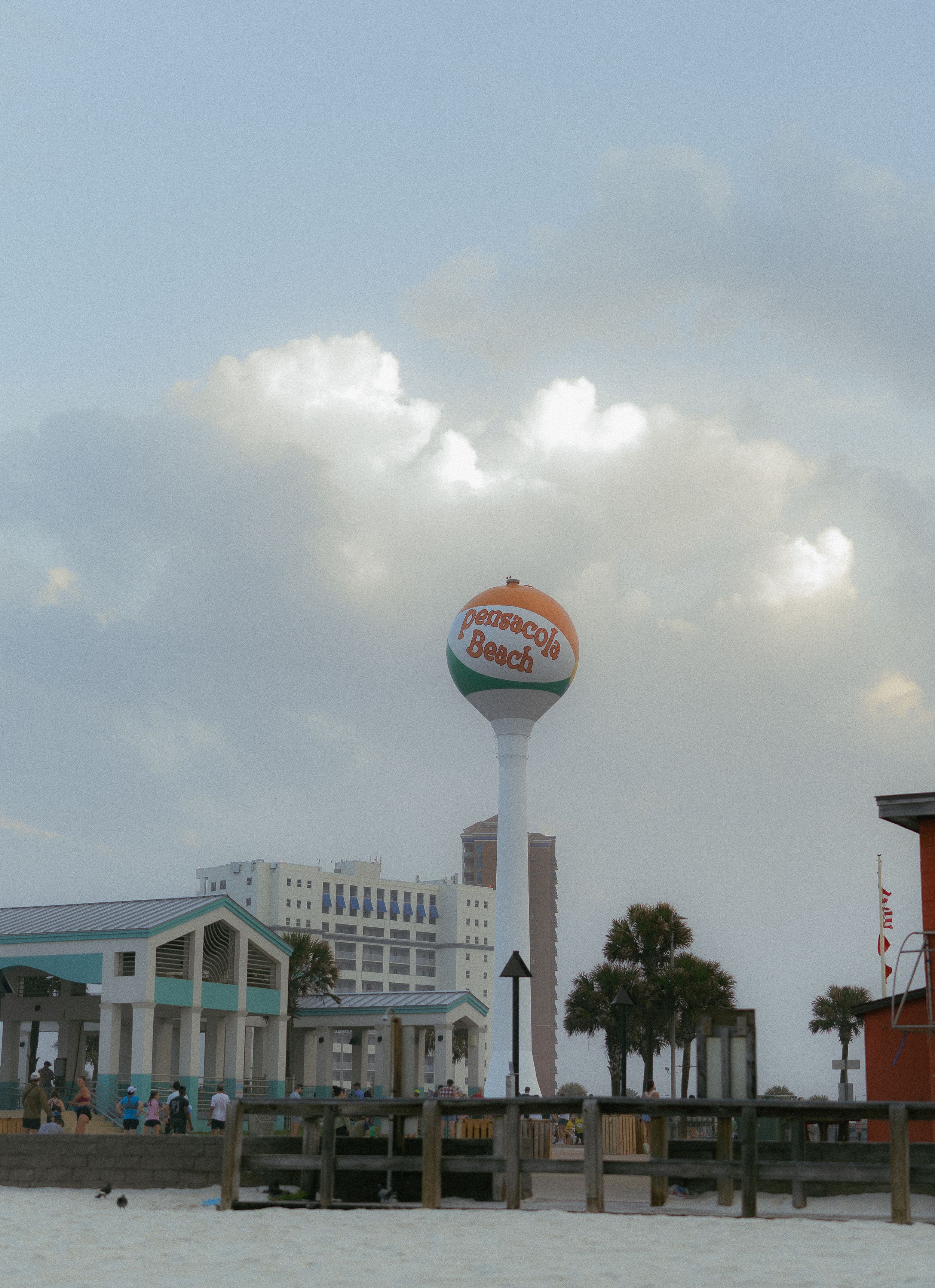A large Pensacola Beach water tower and coastal buildings stand against a cloudy sky along the waterfront.