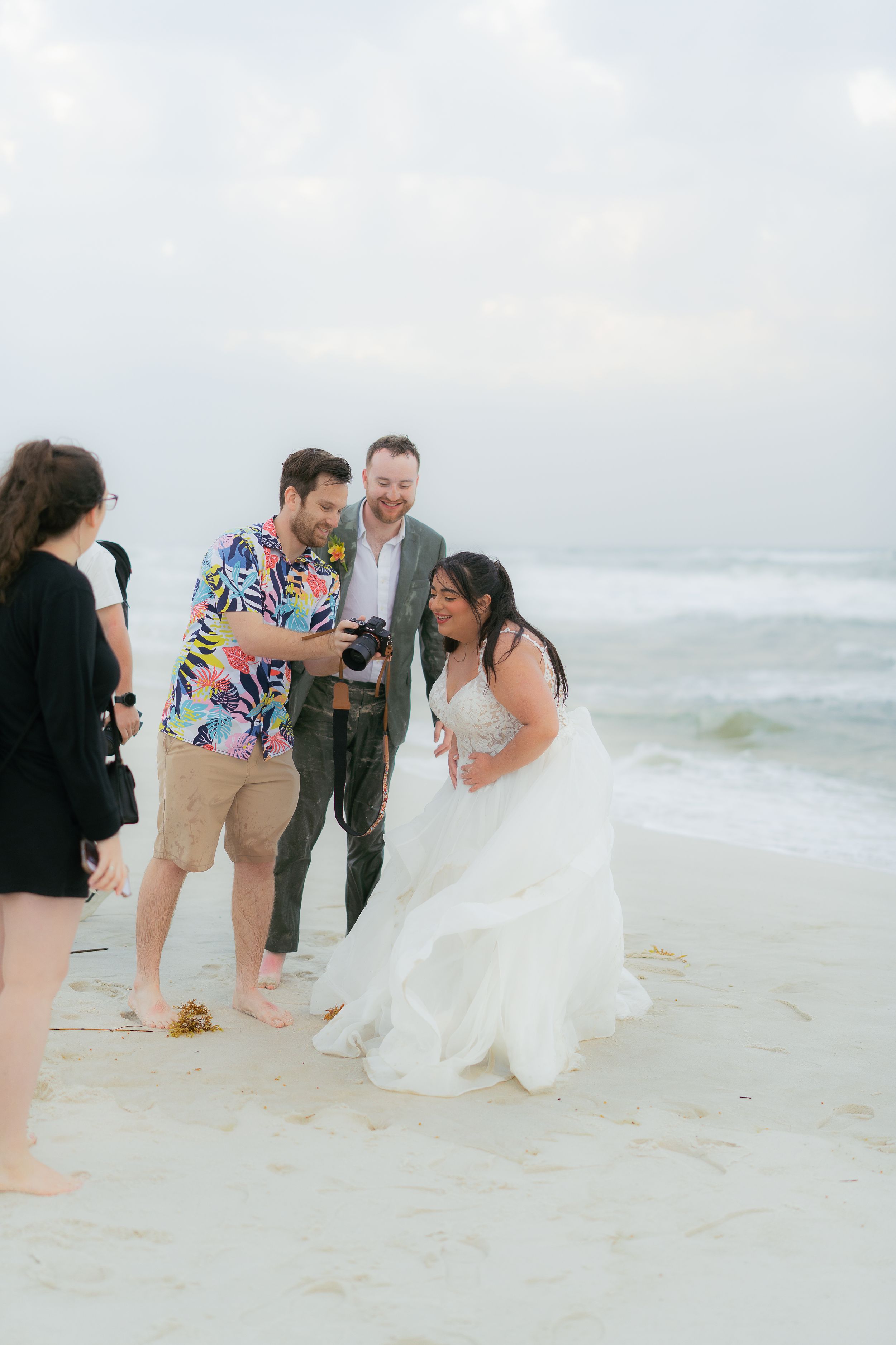 A joyful wedding celebration takes place on a cloudy beach as a group shares a candid moment by the ocean waves.