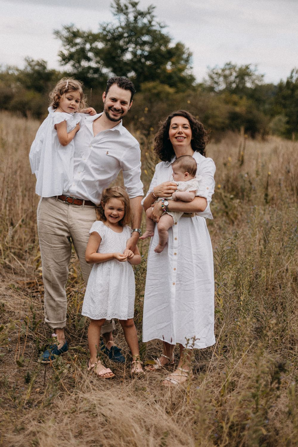 Family of five wearing coordinated white outfits poses together in rustic field setting.