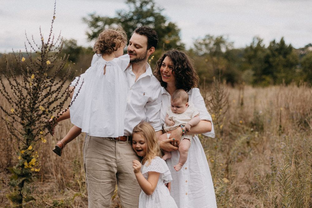 Candid family portrait of parents with three children in white clothing standing in tall grass.