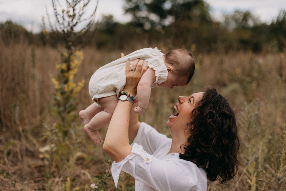 A tender moment captured in a field as a white-clad figure lifts and plays with an infant against a natural backdrop.