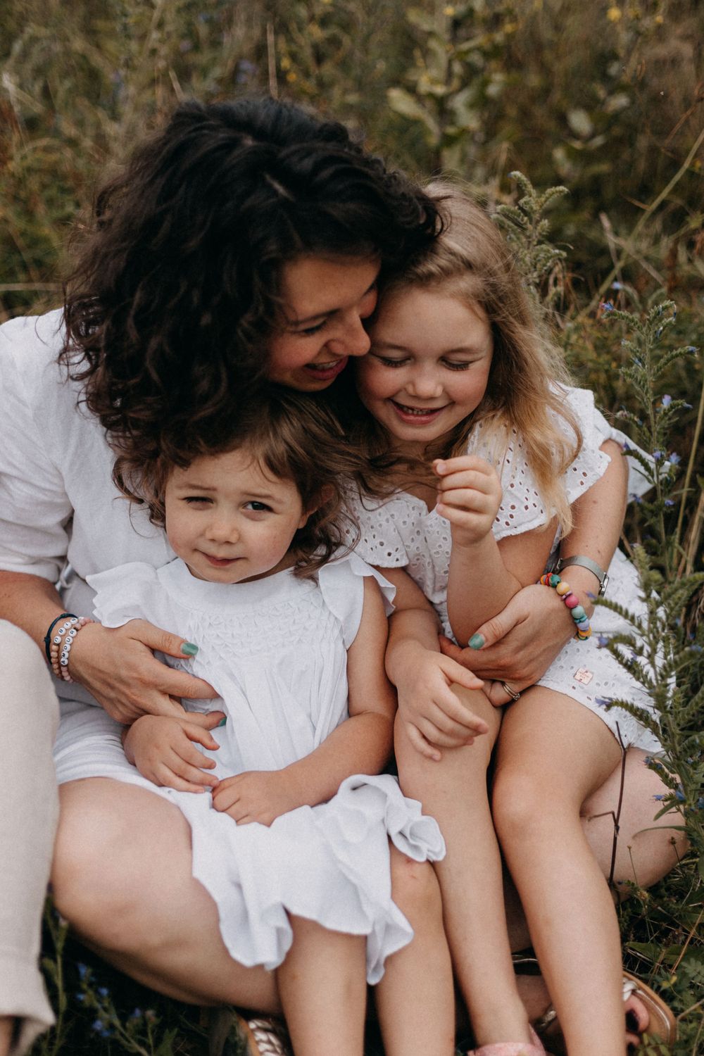 A loving moment captured between parent and children sitting together in a natural outdoor setting wearing white outfits.