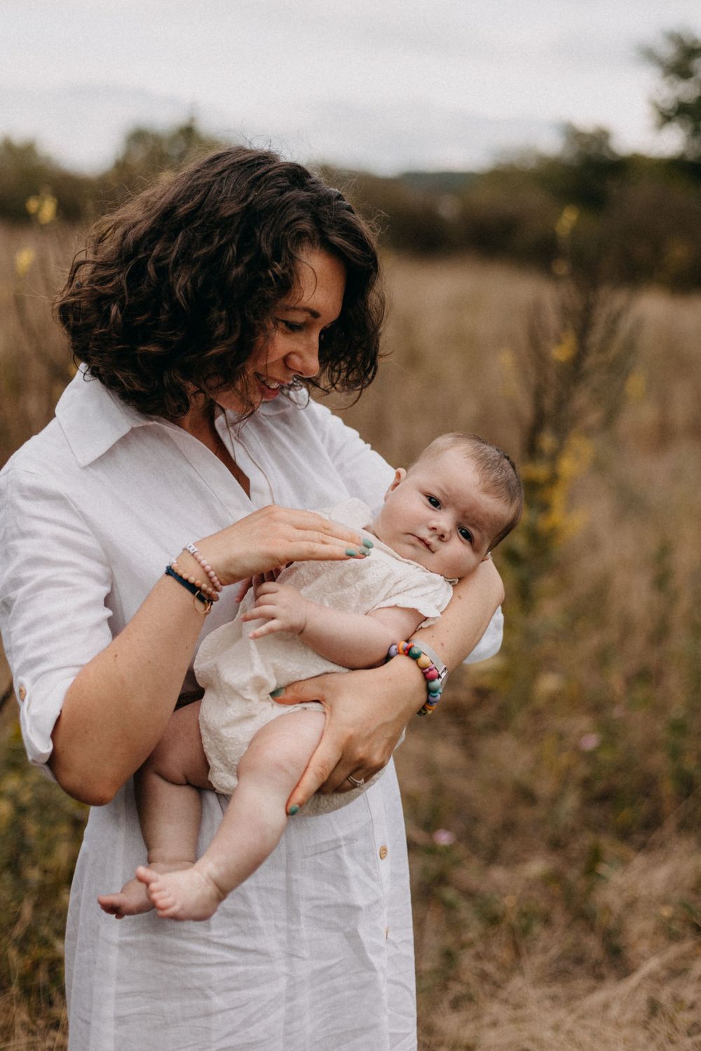 Parent in white dress tenderly holding and looking down at infant in outdoor field setting.