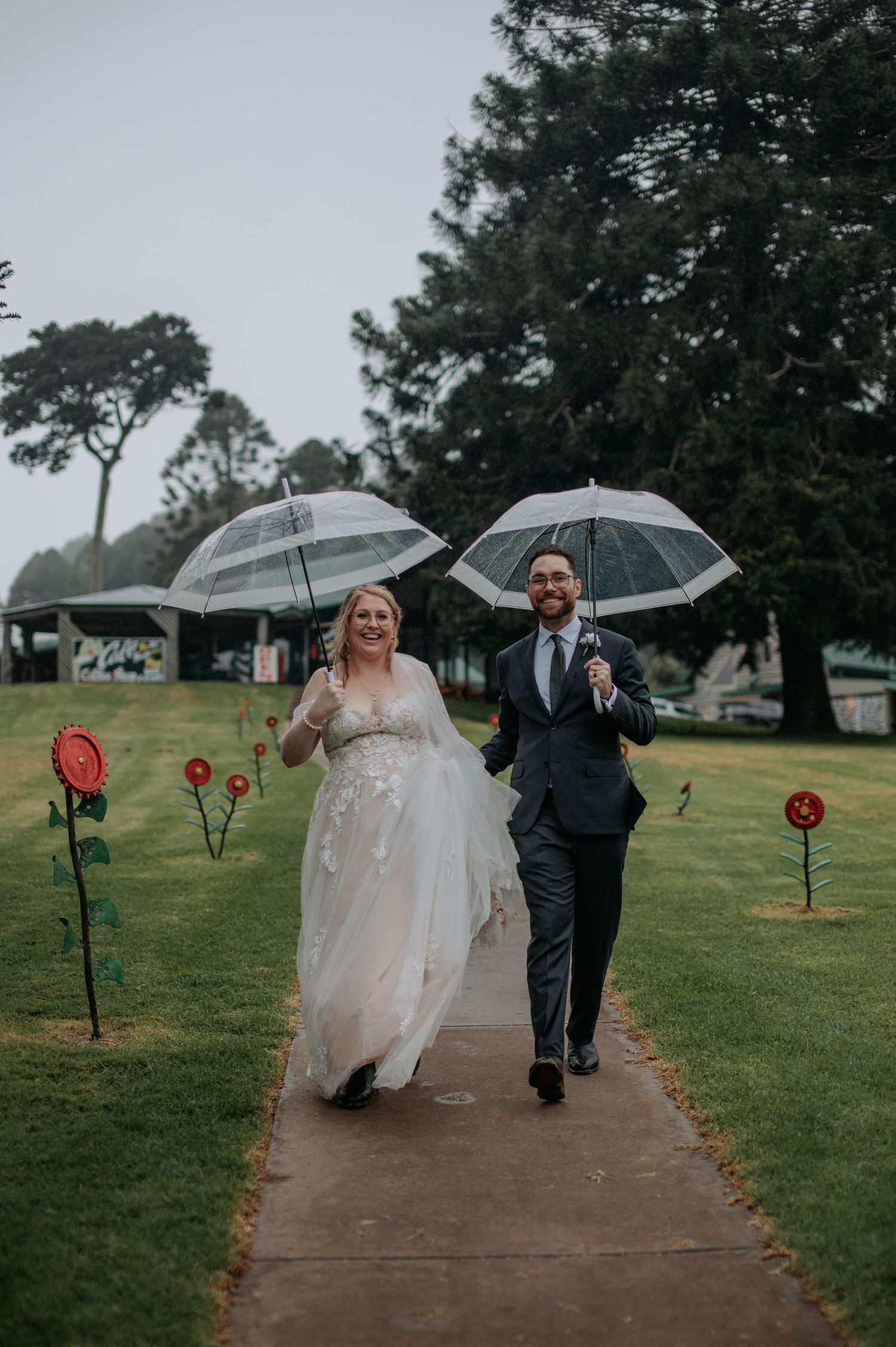 Couple walking with clear umbrellas on rainy wedding day past decorative red flowers.