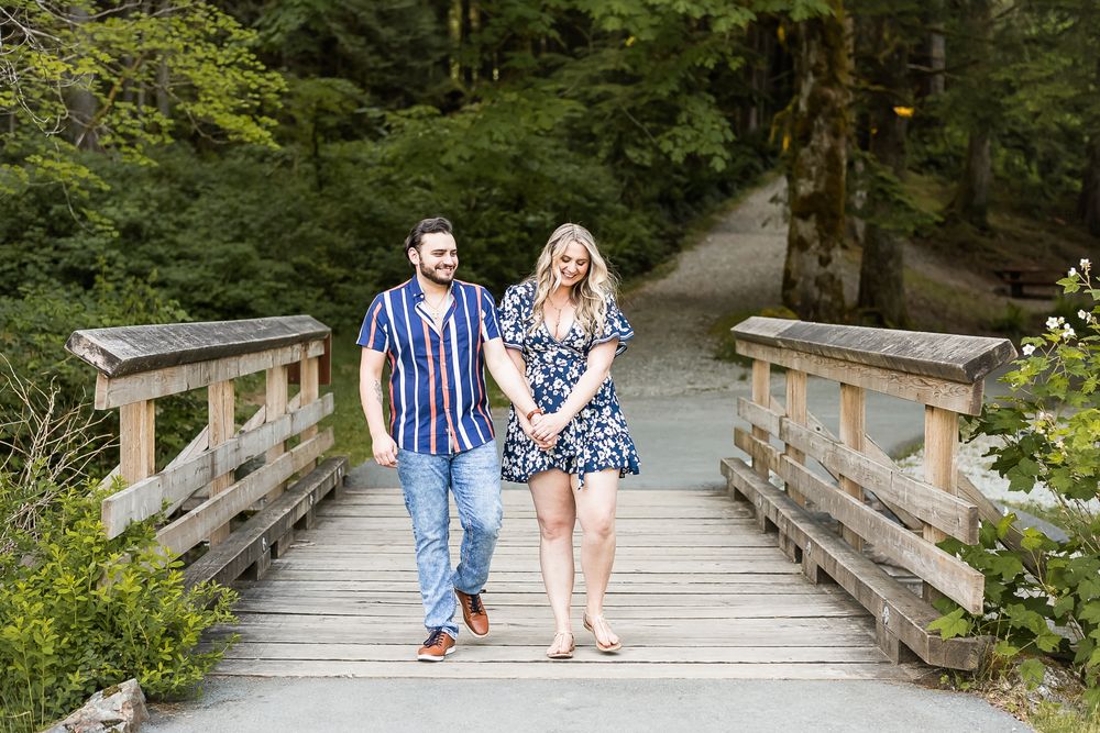 Holding hands and walking together over a wooden bridge in the forest, a couple smiles and laughs.