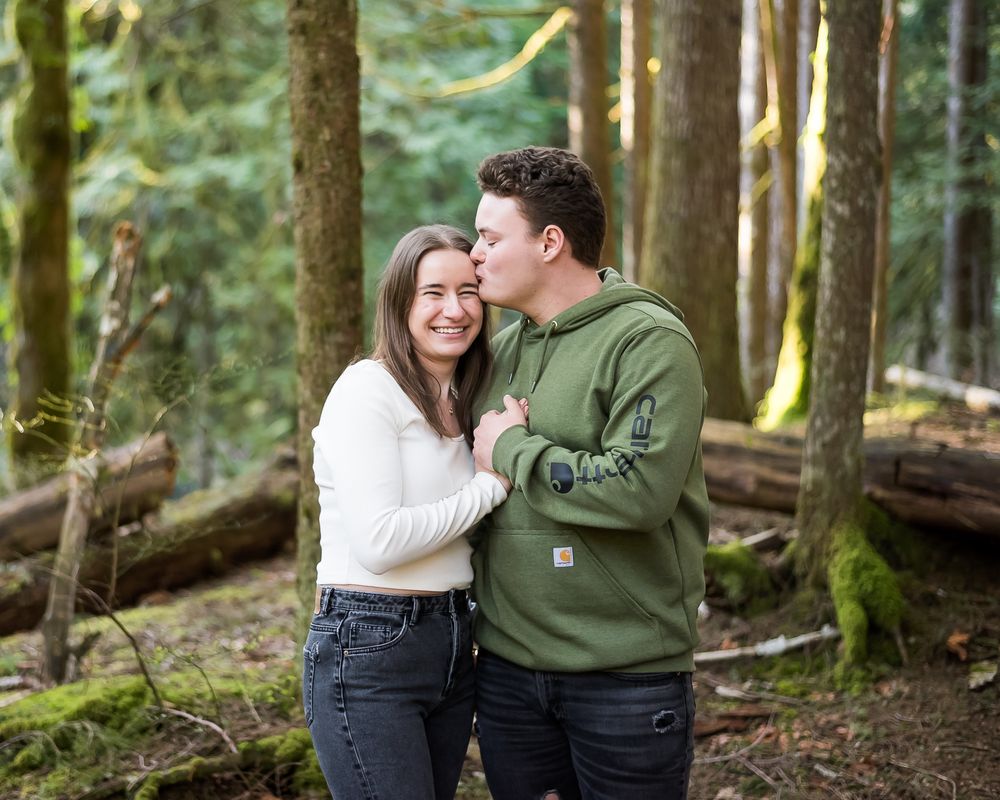 A man kisses his fiancee on the cheek during their engagement session in a pacific northwest forest.