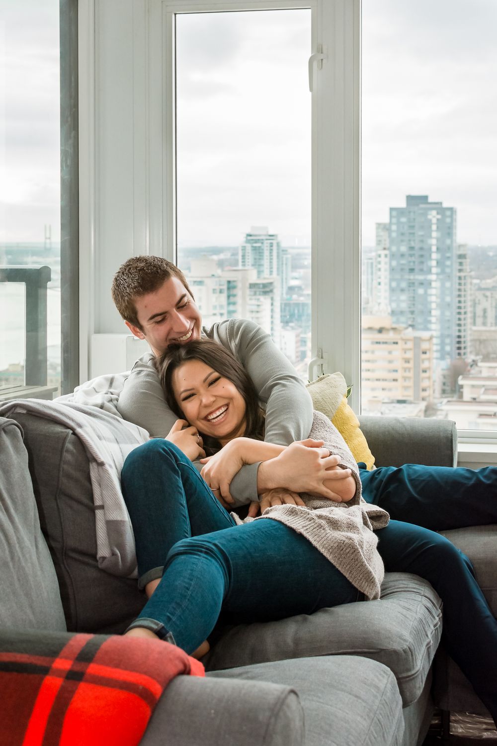 A couple cuddle up together on the couch and smile at the camera as their celebrate their engagement.