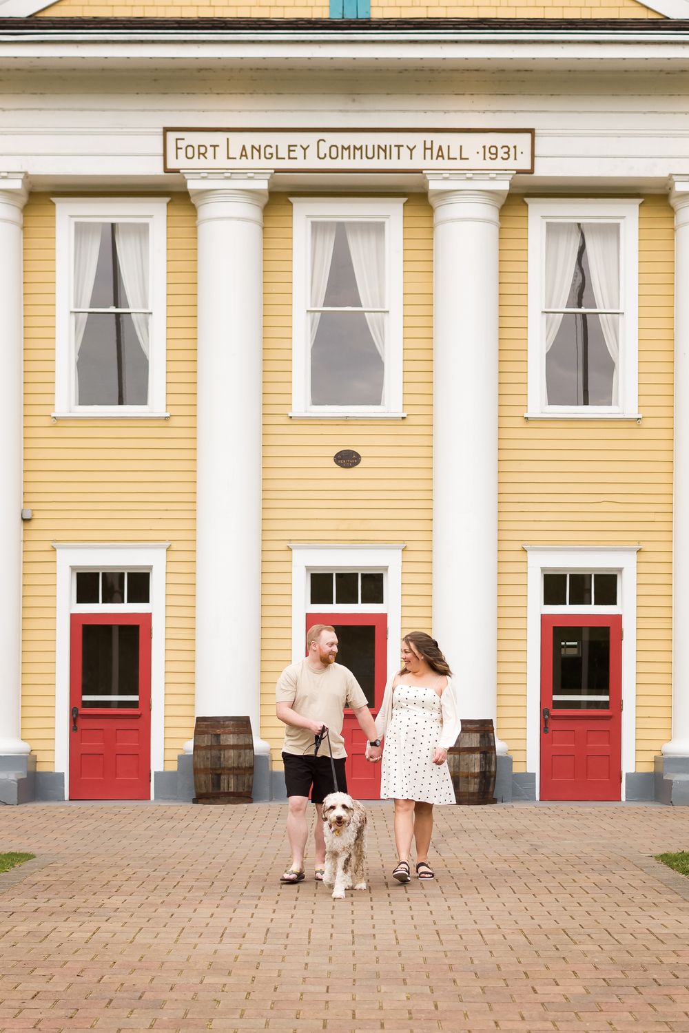 A couple hold hands and walk together with their dog in front of the Fort Langley Community Hall.