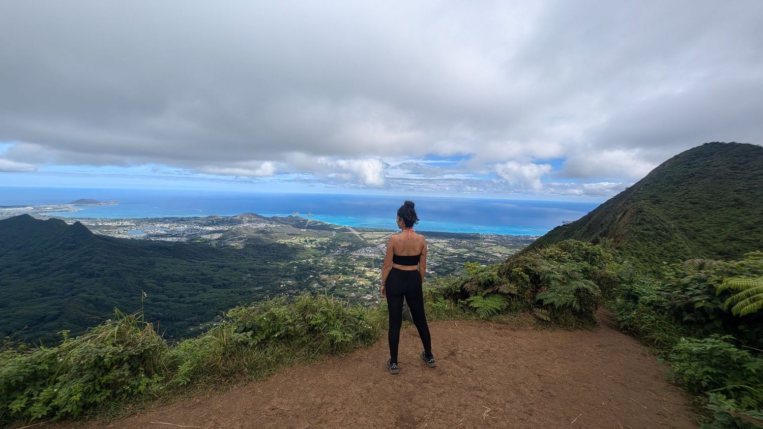 Woman standing on a scenic mountain overlook in Hawaii, gazing at lush green valleys and ocean views under cloudy skies.