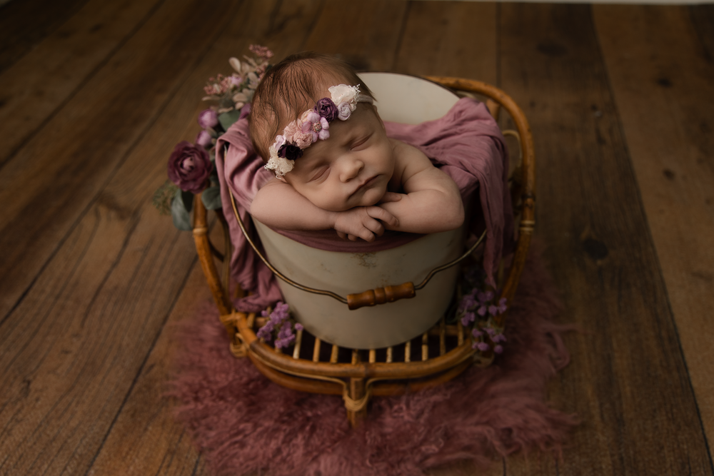 Newborn baby sleeping peacefully in a wicker bucket with pink floral headband and soft mauve blanket on wood floor.