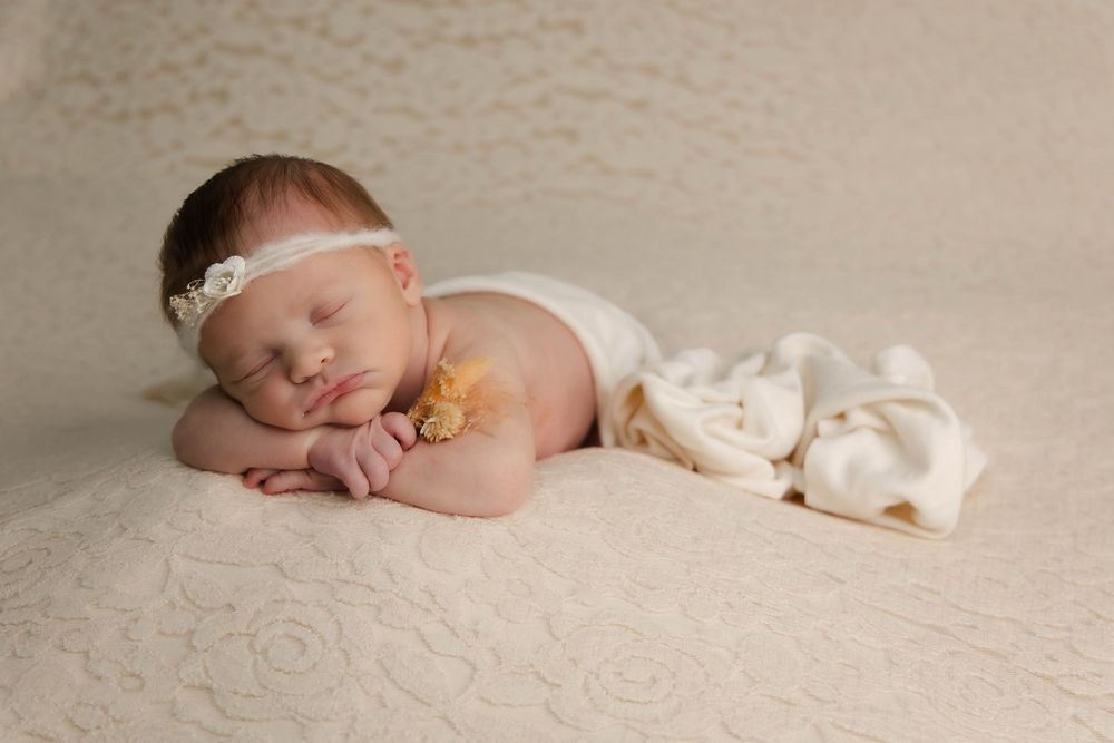 Peaceful newborn baby sleeping on cream lace blanket wearing white headband.