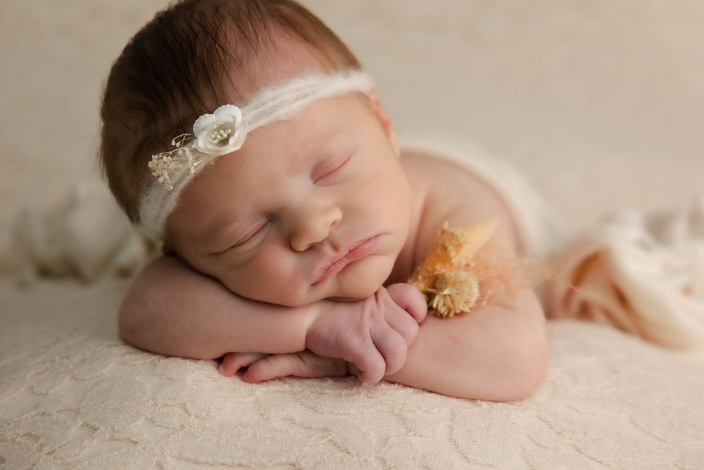 Sleeping newborn baby girl wearing a white floral headband, resting on hands on a cream lace blanket.