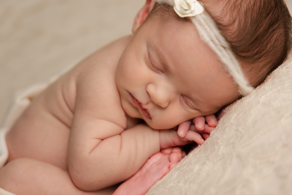 Peaceful newborn baby girl sleeping with hands tucked under chin, wearing a delicate white floral headband.