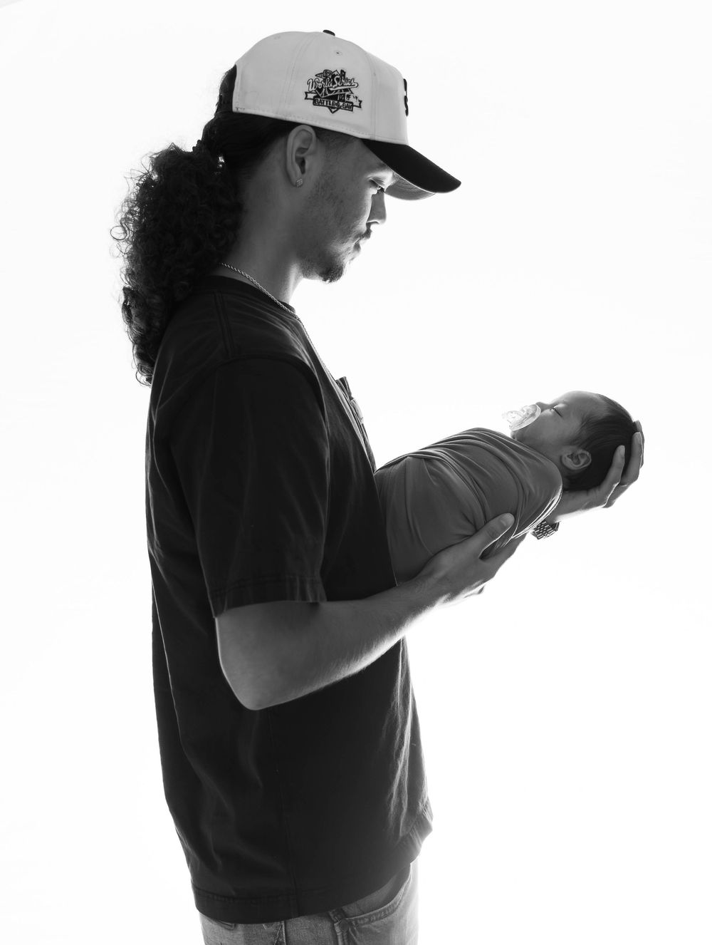 Black and white photo of a man with long hair and trucker hat tenderly holding a newborn baby.