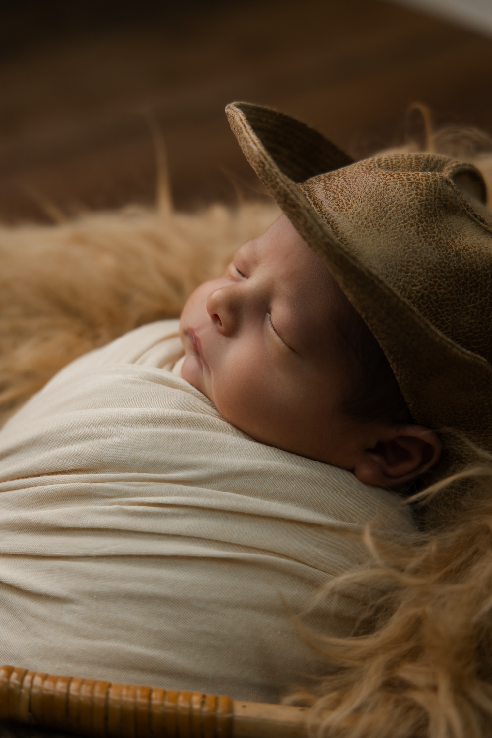 Sleeping newborn baby wearing an oversized brown cowboy hat, wrapped in cream swaddle cloth in a wicker basket.