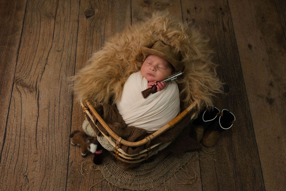 Newborn baby swaddled in white wrap sleeping in a wicker basket with fur lining, wearing a tiny hat with a bow tie prop.